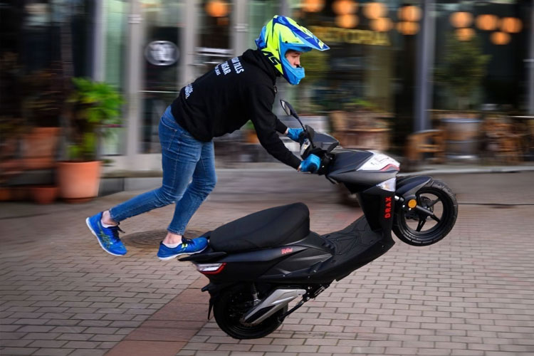 En ung man som står med ena foten på en moped som kör på bakhjulet i cirklar